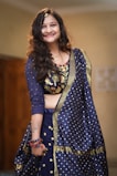 A person wearing a traditional blue attire with intricate embroidery and polka dot patterns. The clothing includes a richly decorated blouse and matching dupatta. The person is smiling and accessorized with bracelets and a hair ornament. There is a tattoo visible on the arm.