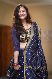 A person wearing a traditional blue attire with intricate embroidery and polka dot patterns. The clothing includes a richly decorated blouse and matching dupatta. The person is smiling and accessorized with bracelets and a hair ornament. There is a tattoo visible on the arm.