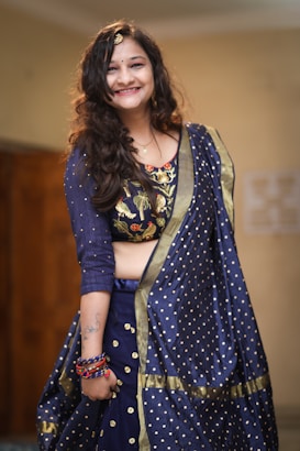 A person wearing a traditional blue attire with intricate embroidery and polka dot patterns. The clothing includes a richly decorated blouse and matching dupatta. The person is smiling and accessorized with bracelets and a hair ornament. There is a tattoo visible on the arm.