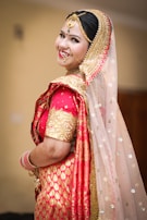 A person dressed in traditional Indian bridal attire with elaborate jewelry, including a nose ring and a headpiece. The attire features vibrant red and gold colors with intricate patterns and embellishments. The veil is sheer with sparkling details.