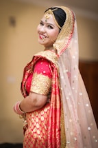 A person dressed in traditional Indian bridal attire with elaborate jewelry, including a nose ring and a headpiece. The attire features vibrant red and gold colors with intricate patterns and embellishments. The veil is sheer with sparkling details.