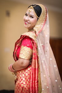 A person dressed in traditional Indian bridal attire with elaborate jewelry, including a nose ring and a headpiece. The attire features vibrant red and gold colors with intricate patterns and embellishments. The veil is sheer with sparkling details.