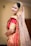 A person dressed in traditional Indian bridal attire with elaborate jewelry, including a nose ring and a headpiece. The attire features vibrant red and gold colors with intricate patterns and embellishments. The veil is sheer with sparkling details.