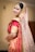 A person dressed in traditional Indian bridal attire with elaborate jewelry, including a nose ring and a headpiece. The attire features vibrant red and gold colors with intricate patterns and embellishments. The veil is sheer with sparkling details.
