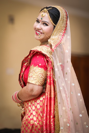A person dressed in traditional Indian bridal attire with elaborate jewelry, including a nose ring and a headpiece. The attire features vibrant red and gold colors with intricate patterns and embellishments. The veil is sheer with sparkling details.