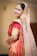A person dressed in traditional Indian bridal attire with elaborate jewelry, including a nose ring and a headpiece. The attire features vibrant red and gold colors with intricate patterns and embellishments. The veil is sheer with sparkling details.