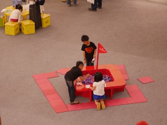 Three children are playing around a red activity table filled with colorful items, likely toys or craft materials. They are situated on a gray floor, and the table is surrounded by red floor mats. Nearby, a couple of small yellow tables with children and adults are visible.
