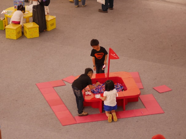 Three children are playing around a red activity table filled with colorful items, likely toys or craft materials. They are situated on a gray floor, and the table is surrounded by red floor mats. Nearby, a couple of small yellow tables with children and adults are visible.