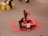 Three children are playing around a red activity table filled with colorful items, likely toys or craft materials. They are situated on a gray floor, and the table is surrounded by red floor mats. Nearby, a couple of small yellow tables with children and adults are visible.