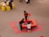 Three children are playing around a red activity table filled with colorful items, likely toys or craft materials. They are situated on a gray floor, and the table is surrounded by red floor mats. Nearby, a couple of small yellow tables with children and adults are visible.