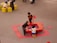 Three children are playing around a red activity table filled with colorful items, likely toys or craft materials. They are situated on a gray floor, and the table is surrounded by red floor mats. Nearby, a couple of small yellow tables with children and adults are visible.