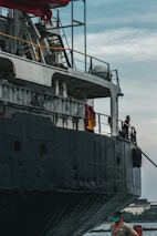 A large ship with a dark exterior is docked alongside a body of water. The ship is equipped with many structural elements and railings. A worker wearing a safety vest and a mask stands on the dock near the ship.