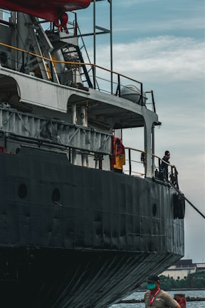 A large ship with a dark exterior is docked alongside a body of water. The ship is equipped with many structural elements and railings. A worker wearing a safety vest and a mask stands on the dock near the ship.