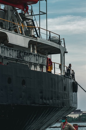 A large ship with a dark exterior is docked alongside a body of water. The ship is equipped with many structural elements and railings. A worker wearing a safety vest and a mask stands on the dock near the ship.