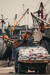 A bustling port scene with a worker in an orange shirt standing atop a truck loaded with bags. Wooden ships with tall masts line the background, and another person can be seen in the distance.