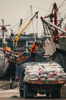 A bustling port scene with a worker in an orange shirt standing atop a truck loaded with bags. Wooden ships with tall masts line the background, and another person can be seen in the distance.