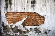A weathered brick wall with a large patch of exposed red bricks surrounded by eroded white plaster. Streaks and stains run vertically down the plaster, suggesting water damage or aging.
