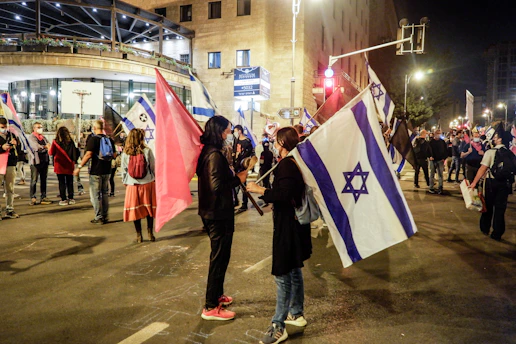 A nighttime protest scene on an urban street with individuals holding flags. Several people are engaged in conversations and many are carrying or waving flags featuring a blue Star of David on a white background, alongside others with pink flags. The street is well-lit with nearby buildings and streetlights visible, creating a bustling, active atmosphere.