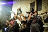 Several individuals are gathered outside a building, holding professional cameras and taking photographs. The group appears focused on capturing an event, with some wearing masks and using flash attachments. The building has visible stone walls and bars on the windows.