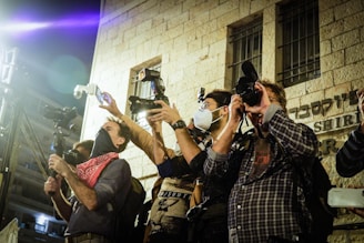 Several individuals are gathered outside a building, holding professional cameras and taking photographs. The group appears focused on capturing an event, with some wearing masks and using flash attachments. The building has visible stone walls and bars on the windows.