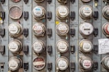 A grid of multiple analog and digital electricity meters mounted on a wall, each with labels and numbers. Some meters have colored seals, and there are handwritten notes and graffiti on the surface. The meters are encased in glass and mounted in a metal frame.