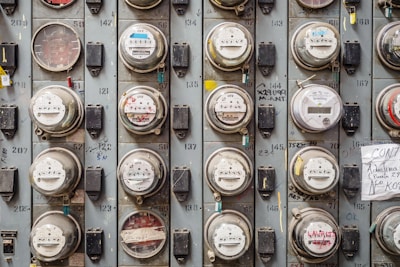 A grid of multiple analog and digital electricity meters mounted on a wall, each with labels and numbers. Some meters have colored seals, and there are handwritten notes and graffiti on the surface. The meters are encased in glass and mounted in a metal frame.