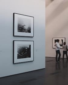 A minimalist art gallery features monochrome photographs on white walls. Two pieces are prominently displayed, with one larger photo below a smaller one. The gallery environment is calm and pristine, with smooth dark flooring and softly lit walls. Two individuals are engaged in viewing the artwork from a distance.