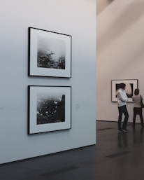 A minimalist art gallery features monochrome photographs on white walls. Two pieces are prominently displayed, with one larger photo below a smaller one. The gallery environment is calm and pristine, with smooth dark flooring and softly lit walls. Two individuals are engaged in viewing the artwork from a distance.