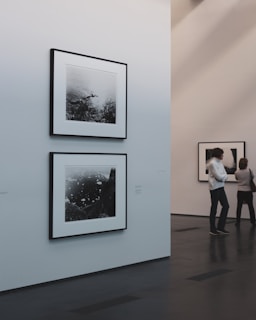 A minimalist art gallery features monochrome photographs on white walls. Two pieces are prominently displayed, with one larger photo below a smaller one. The gallery environment is calm and pristine, with smooth dark flooring and softly lit walls. Two individuals are engaged in viewing the artwork from a distance.