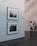 A minimalist art gallery features monochrome photographs on white walls. Two pieces are prominently displayed, with one larger photo below a smaller one. The gallery environment is calm and pristine, with smooth dark flooring and softly lit walls. Two individuals are engaged in viewing the artwork from a distance.