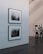 A minimalist art gallery features monochrome photographs on white walls. Two pieces are prominently displayed, with one larger photo below a smaller one. The gallery environment is calm and pristine, with smooth dark flooring and softly lit walls. Two individuals are engaged in viewing the artwork from a distance.