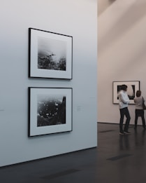 A minimalist art gallery features monochrome photographs on white walls. Two pieces are prominently displayed, with one larger photo below a smaller one. The gallery environment is calm and pristine, with smooth dark flooring and softly lit walls. Two individuals are engaged in viewing the artwork from a distance.
