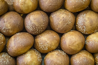 A collection of fresh, round bread rolls with a golden-brown crust, sprinkled with white sesame seeds. The buns are densely arranged, displaying a uniform texture and color, suggesting a freshly baked batch.