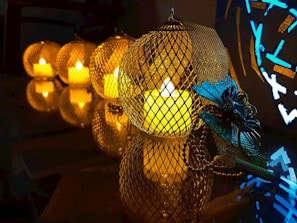 A series of lit candles enclosed in metallic mesh holders create a warm and inviting atmosphere. The setup is arranged on a reflective surface, enhancing the glow and adding depth. A decorative blue butterfly ornament adds a contrasting accent to the scene.