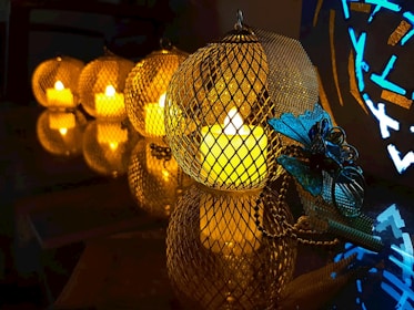 A series of lit candles enclosed in metallic mesh holders create a warm and inviting atmosphere. The setup is arranged on a reflective surface, enhancing the glow and adding depth. A decorative blue butterfly ornament adds a contrasting accent to the scene.