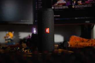 A black cylindrical speaker with a red logo is placed on a wooden surface. In the background, there is a computer monitor displaying a dark interface and various blurred objects, including a figurine and some colorful items.