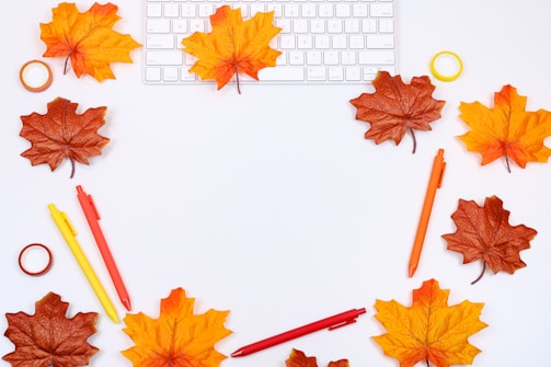 An arrangement of brown and orange autumn leaves surrounds a keyboard, with colorful pens and rolls of tape scattered around. The leaves frame the image against a white background, creating a seasonal theme.