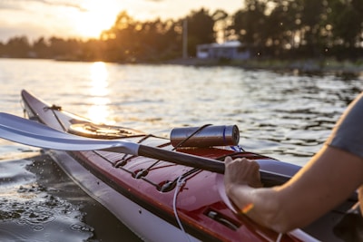 A person is paddling a kayak on a body of water during sunset. The kayak is red and white with a paddle visible. There is a metal thermos attached to the kayak. Trees and buildings line the distant shore, silhouetted by the warm glow of the setting sun.
