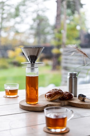 A coffee brewing setup with a glass carafe containing brewed coffee. A metal coffee dripper sits atop the carafe. Nearby, two glass cups filled with coffee are placed on wooden coasters. Next to the setup, there is a pastry on a wooden board, a small metal tin, and a scoop. The background features greenery and trees, suggesting an outdoor or garden setting.