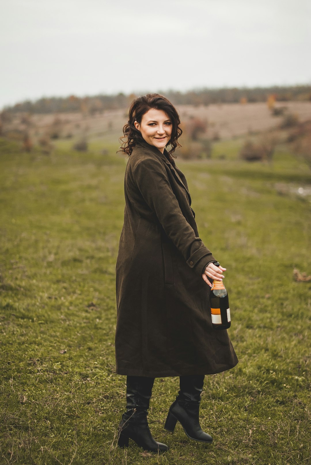 woman in black coat holding bottle standing on green grass field during daytime