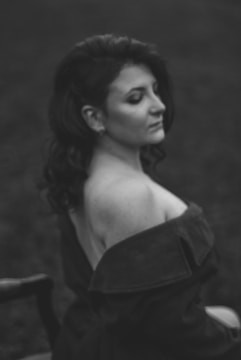 A black and white portrait of a woman with wavy hair sitting on a chair. Her eyes are closed, and she wears an off-shoulder garment. The background is blurred, focusing on her serene expression.