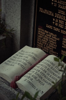 An open book-shaped tombstone with text written in a language that appears to be Indonesian. The headstone behind it contains golden inscriptions with names and dates, indicating it is a memorial site. A red cord is draped across the book, providing a striking contrast to the stone. Surrounding foliage adds a touch of nature to the scene, and the lighting casts a somber, reflective mood.