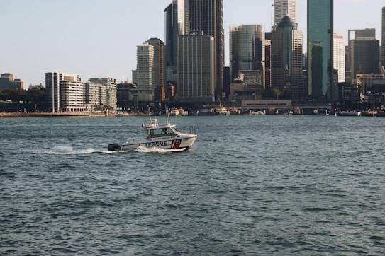 A rescue boat is traveling across the water with a city skyline in the background. Large skyscrapers and various buildings line the shoreline under a clear sky.