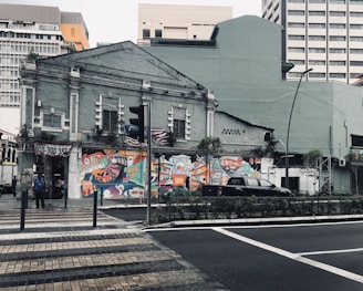 An old building with a faded exterior is decorated with vibrant street art. The artwork features bold colors and abstract designs, adding a splash of creativity to the surroundings. The scene includes a couple of people standing nearby and a car parked on the street. The background shows a mix of modern and old architecture.