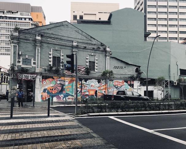 An old building with a faded exterior is decorated with vibrant street art. The artwork features bold colors and abstract designs, adding a splash of creativity to the surroundings. The scene includes a couple of people standing nearby and a car parked on the street. The background shows a mix of modern and old architecture.