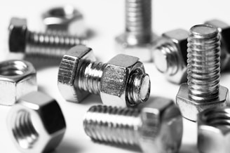 A close-up image featuring several metallic bolts and nuts with a shiny surface, arranged in a scattered manner. The focus highlights the detailed threading of the screws and the hexagonal shape of the nuts.