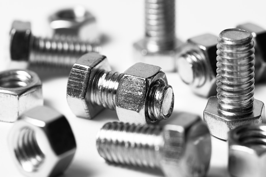 A close-up image featuring several metallic bolts and nuts with a shiny surface, arranged in a scattered manner. The focus highlights the detailed threading of the screws and the hexagonal shape of the nuts.