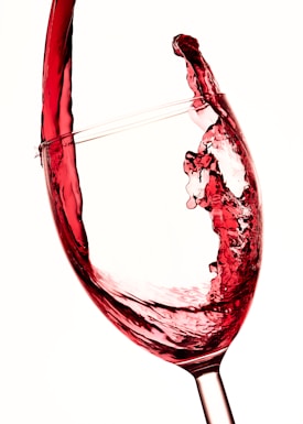 A close-up image of a glass of red wine in motion, with the liquid swirling elegantly inside. The wine is captured mid-splash, revealing the dynamic textures and fluidity. The background is a light, neutral color, which highlights the deep red hue of the wine.