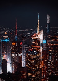 A nighttime cityscape featuring illuminated skyscrapers with distinct architectural designs. The buildings display vibrant lights and advertisements from companies like H&M and Salesforce. The atmosphere is urban and bustling, with a dark sky providing contrast to the bright city lights.