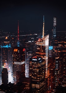 A nighttime cityscape featuring illuminated skyscrapers with distinct architectural designs. The buildings display vibrant lights and advertisements from companies like H&M and Salesforce. The atmosphere is urban and bustling, with a dark sky providing contrast to the bright city lights.